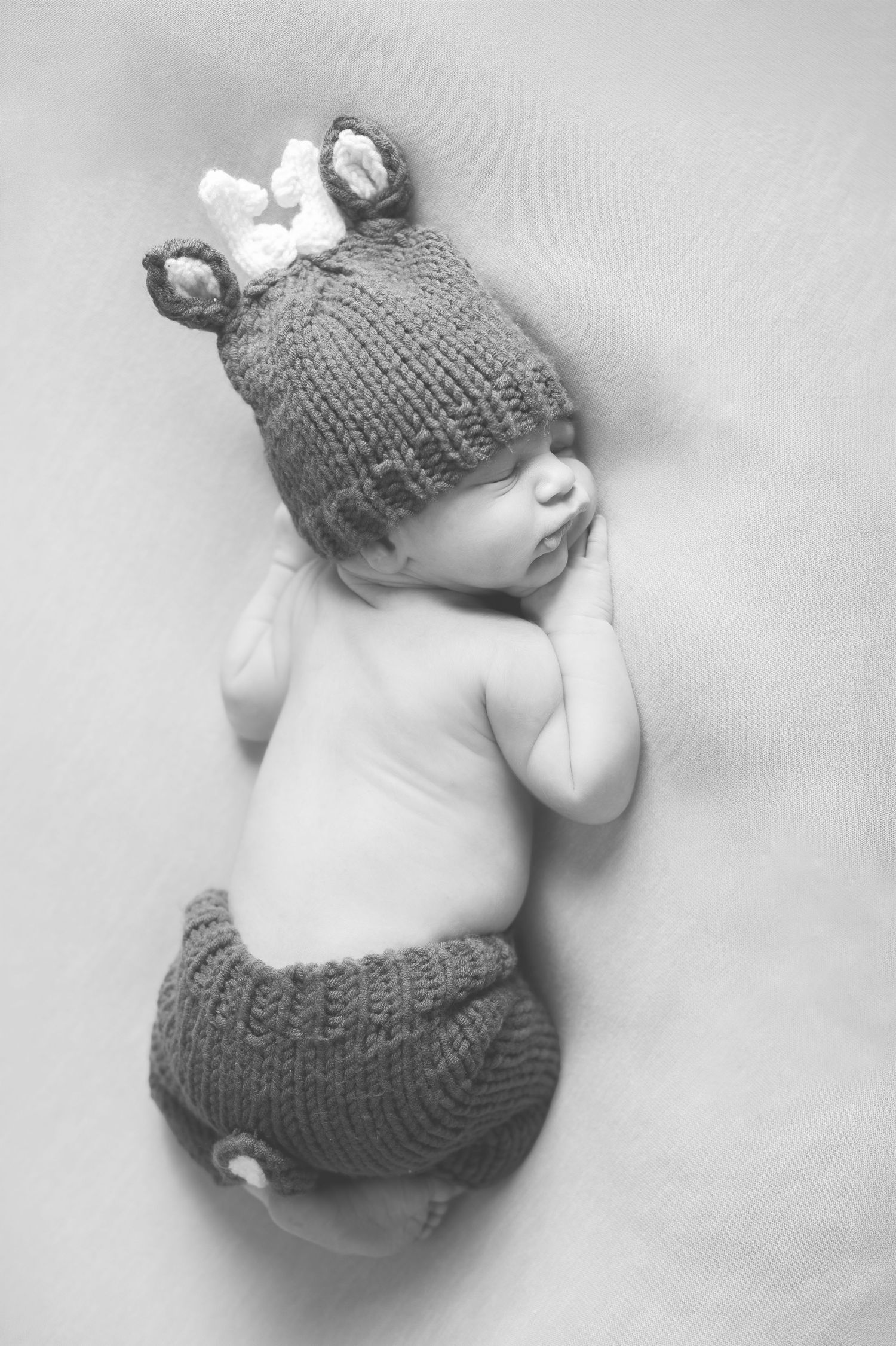 Black and white photo of a sleeping newborn baby wearing a knitted reindeer hat and diaper cover.