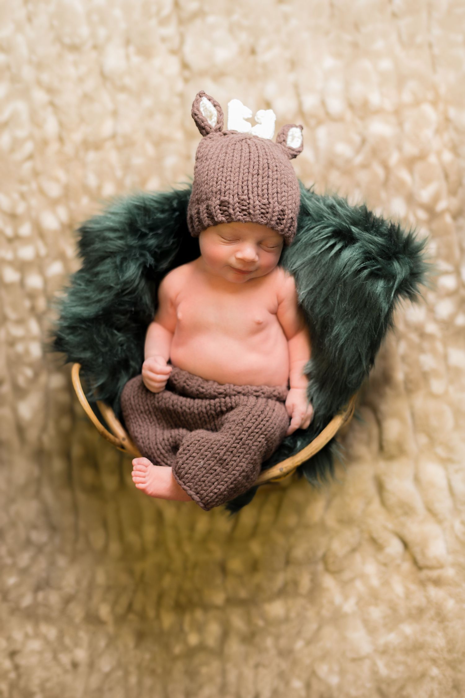 Sleeping newborn baby in a basket wearing a brown knit deer hat and pants on a fluffy green fur blanket.