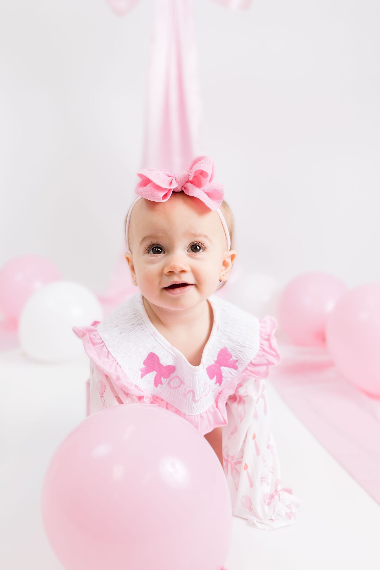 Sweet baby photoshoot with pink bows and soft lighting against white background.