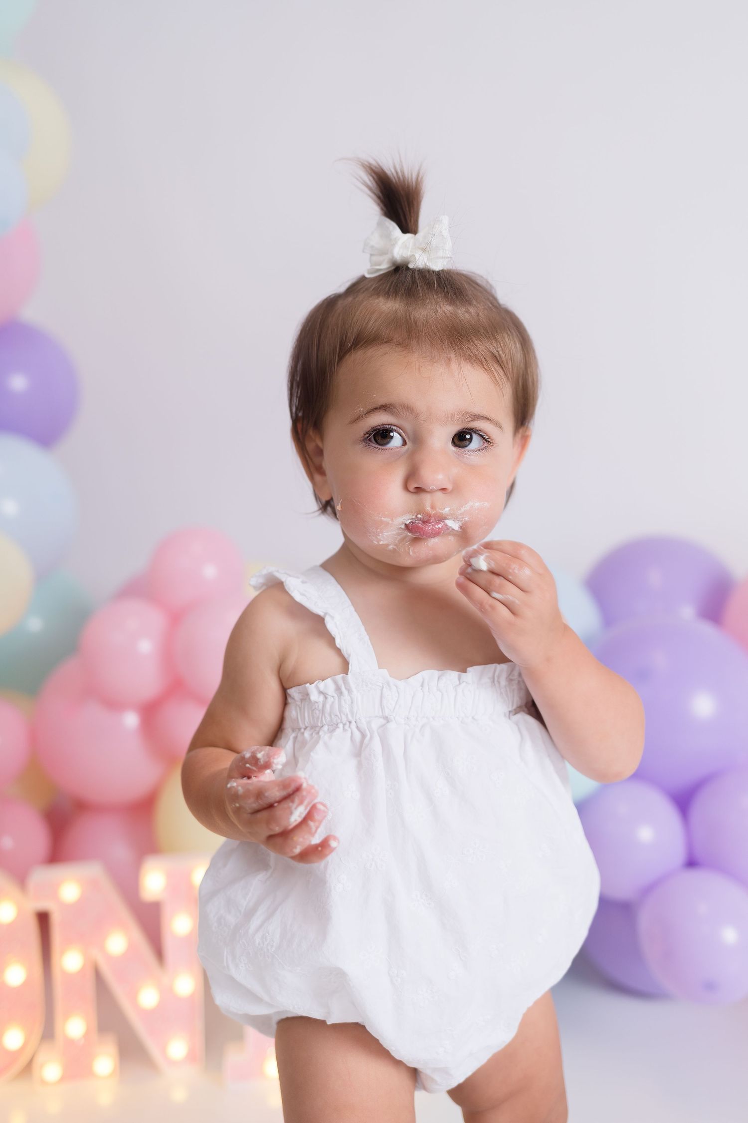 Baby in white outfit with cake-covered face against pastel balloon backdrop.