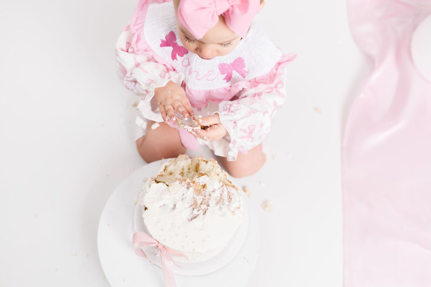 Baby wearing pink dress sits on white blanket surrounded by smashed pink and white birthday cake and frosting.