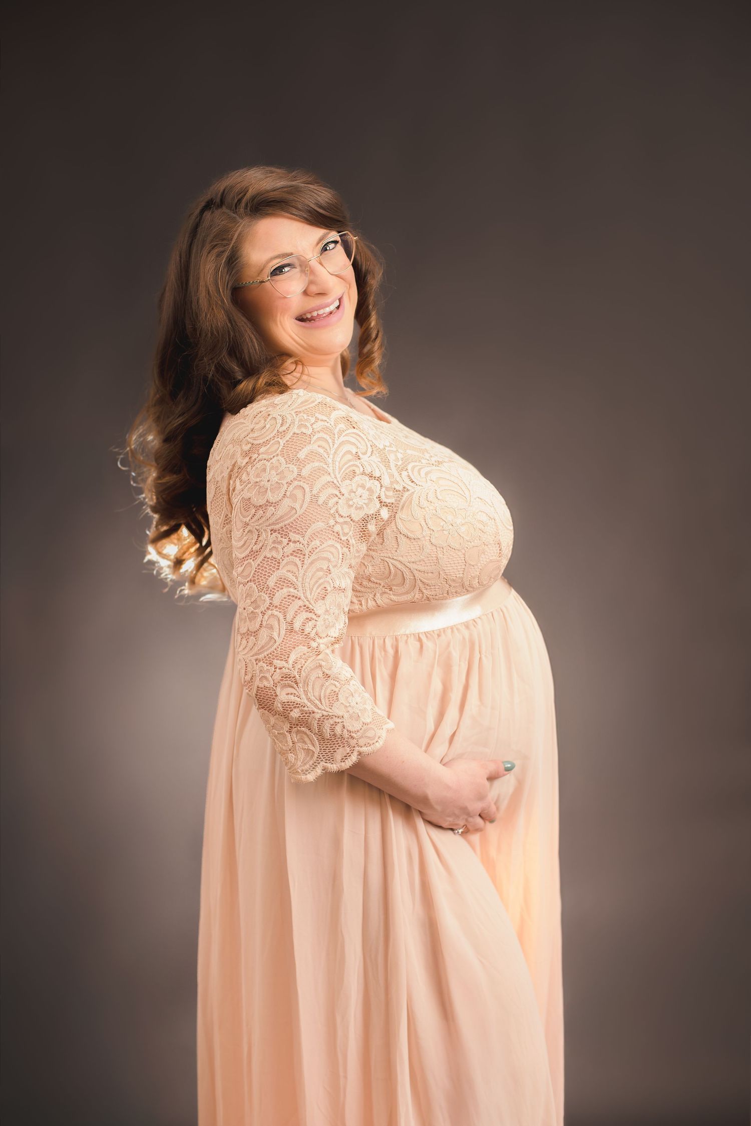 Maternity portrait in flowing peach lace gown against dark studio background.