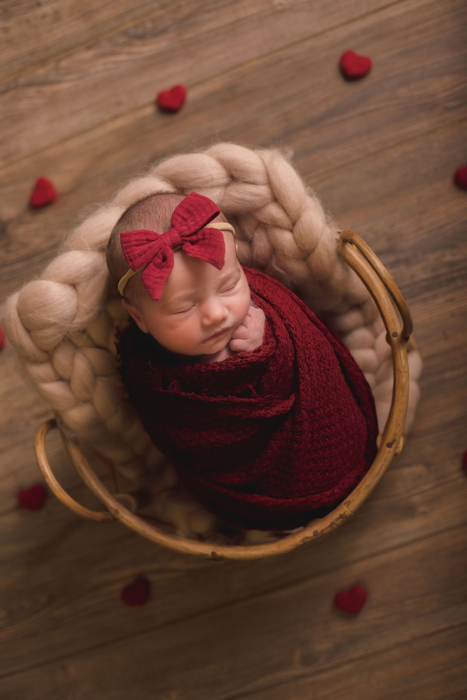 Newborn photography session with red knit wrap and scattered rose petals on wooden floor.