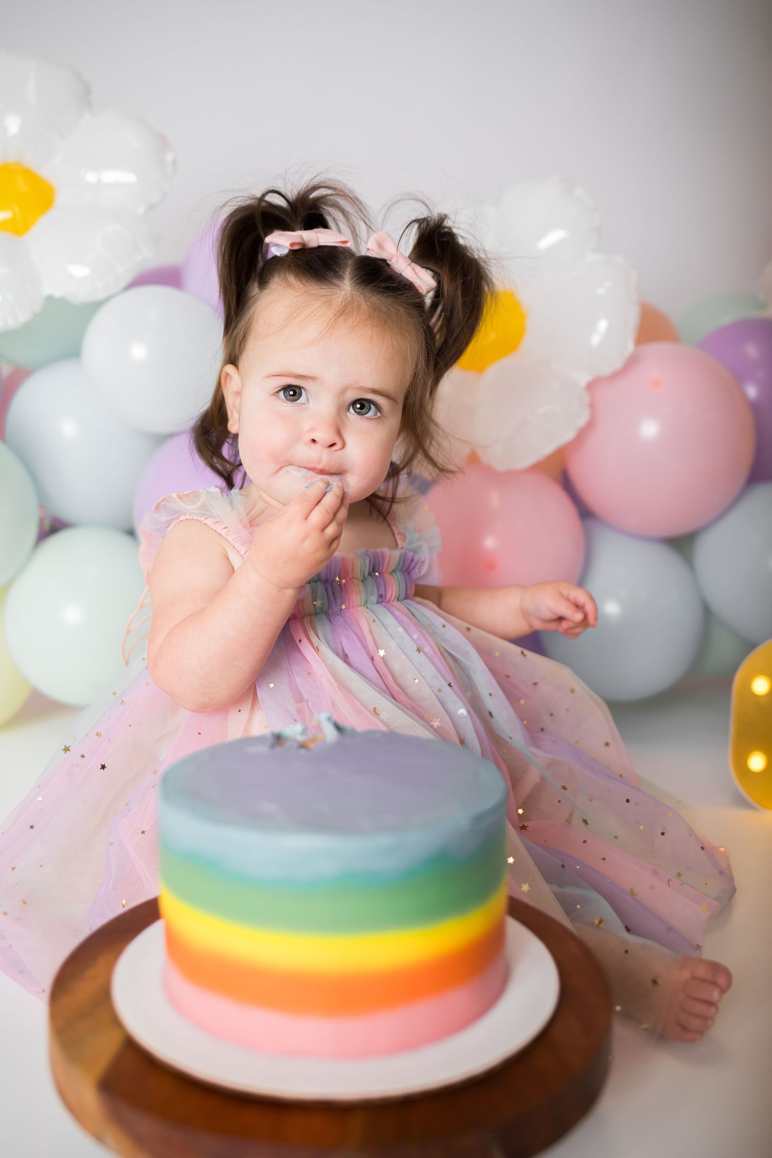 Rainbow layered cake with pastel colors on wooden board surrounded by balloons.