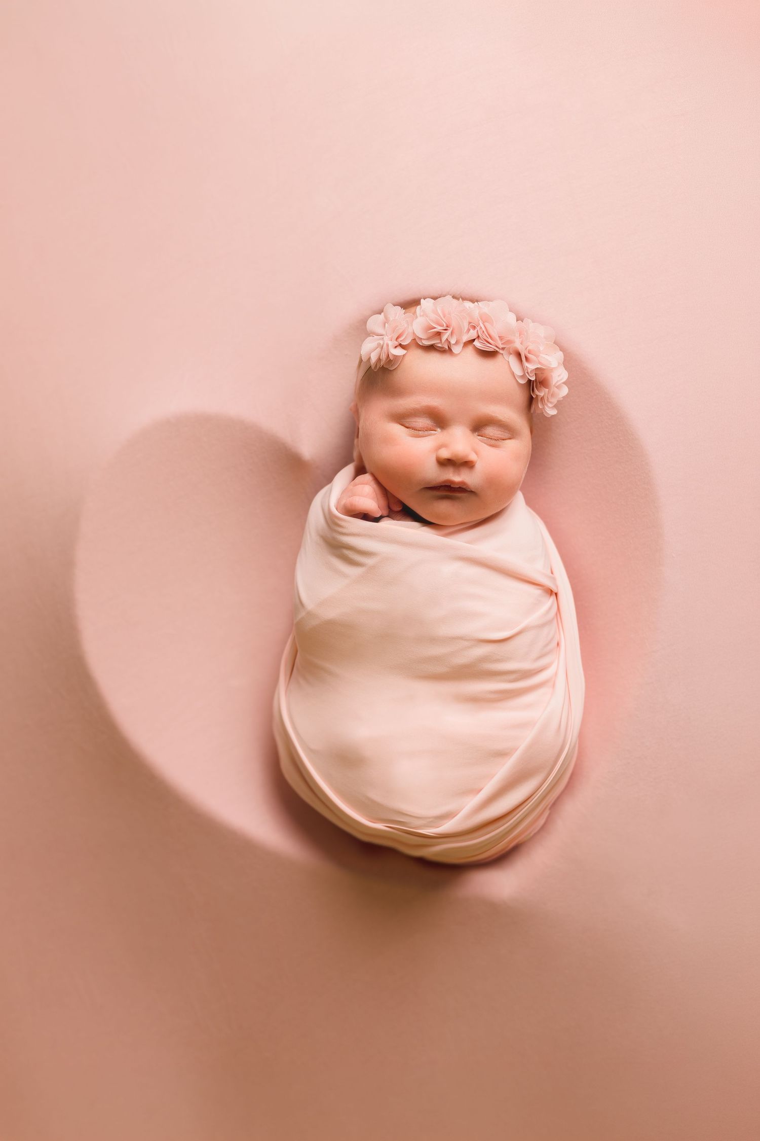 Sleeping newborn baby girl swaddled in cream wrap with pink floral headband on pink heart-shaped background.
