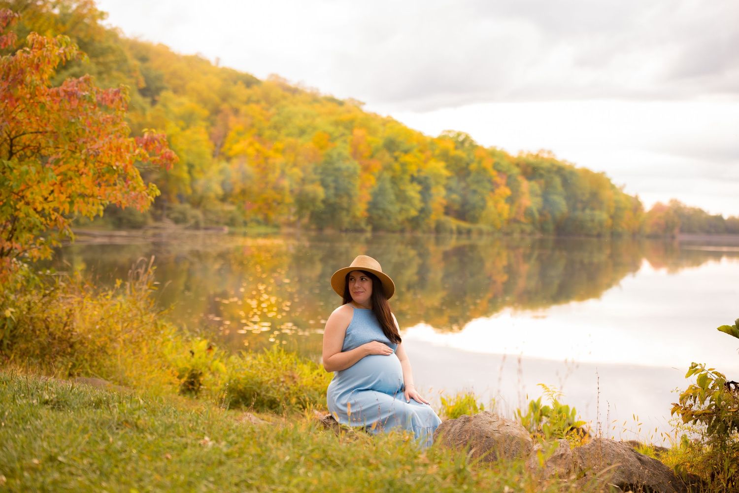Fall maternity session by lakeside with colorful autumn foliage reflecting on water.