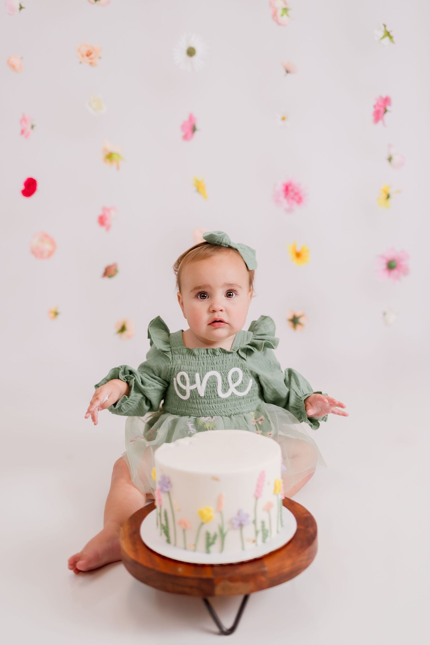 Baby in sage green dress sits with white birthday cake against floral backdrop.