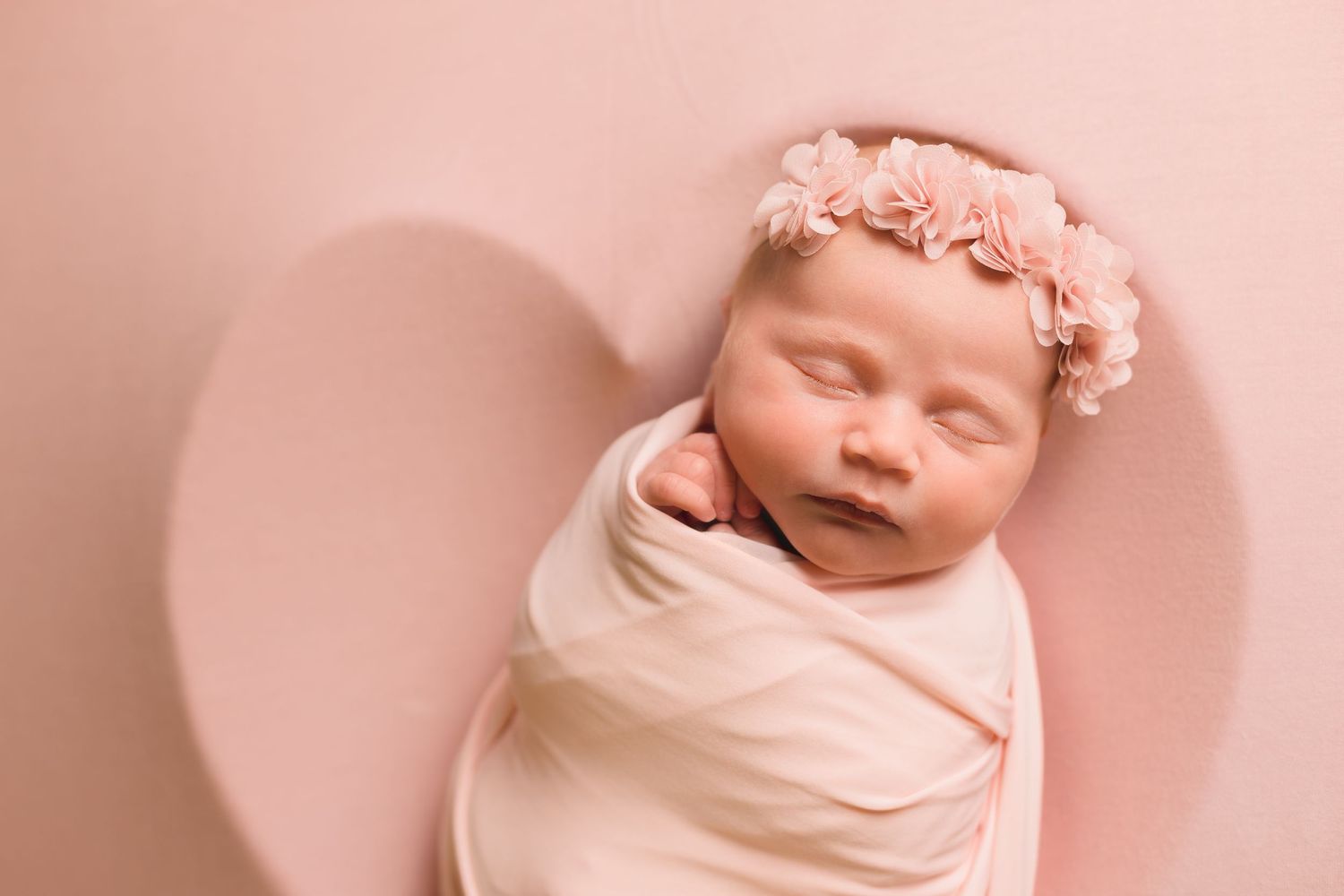 Sleeping newborn baby girl swaddled in pink wrap wearing floral headband on pink heart background.