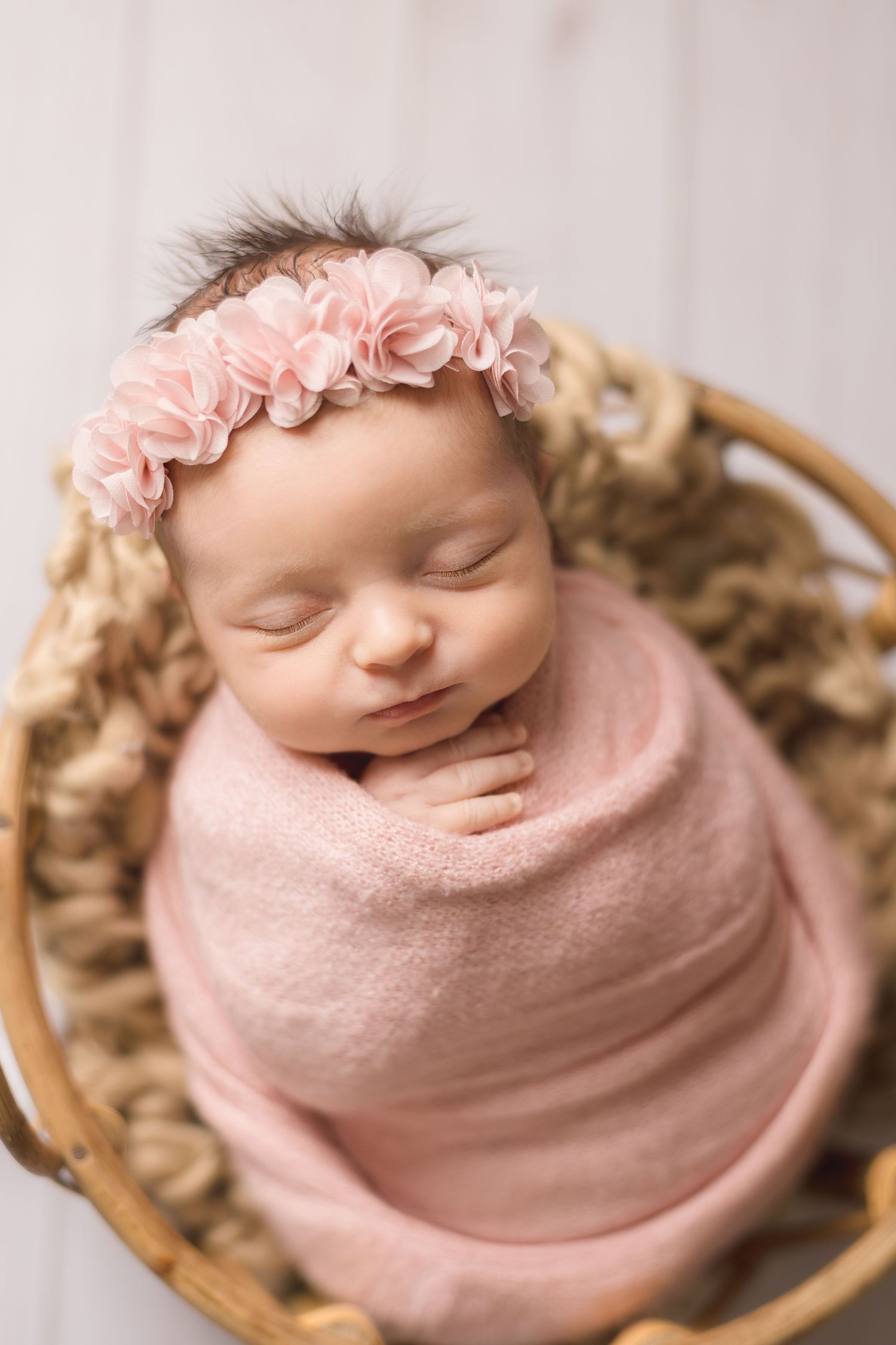 A sleeping newborn baby wrapped in soft pink fabric rests peacefully in a wooden bowl against a neutral background.