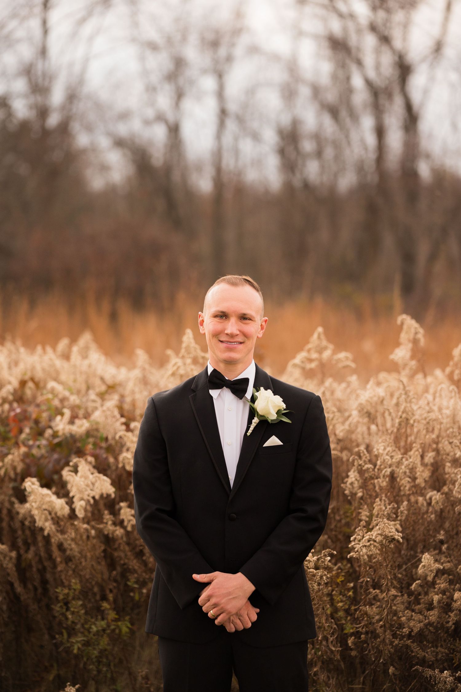 Groom in classic black tuxedo with boutonniere poses in front of golden autumn field at sunset.