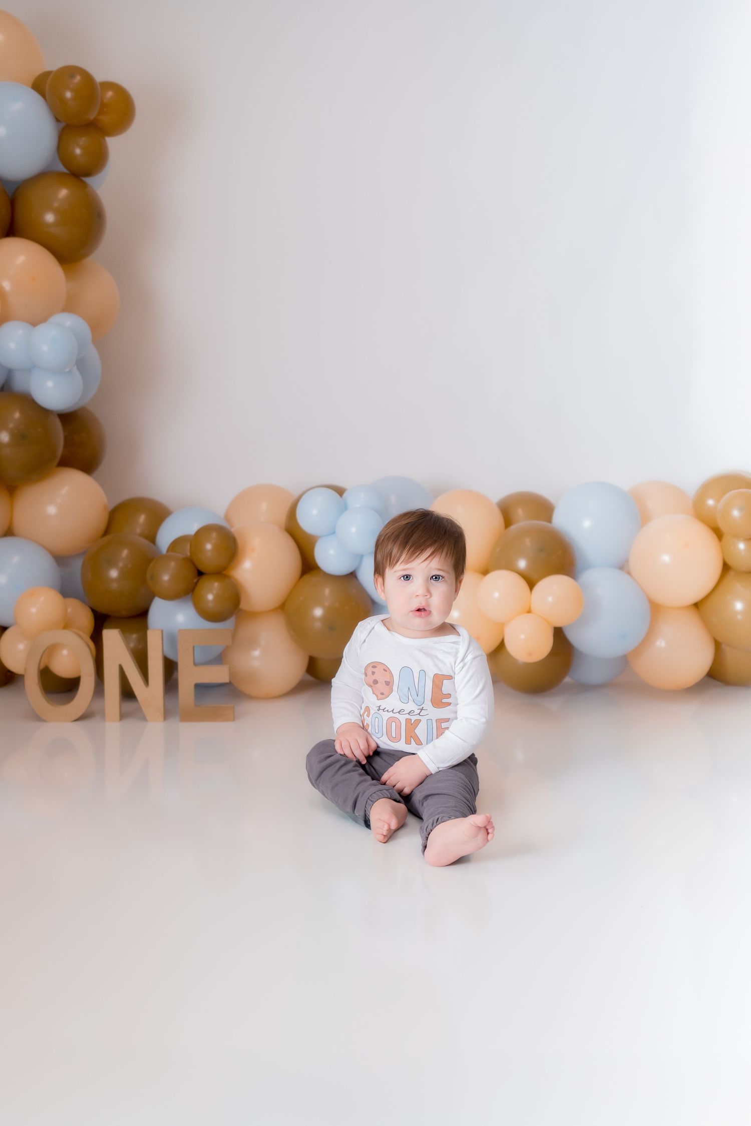 First birthday celebration setup with brown, peach and blue balloon garland against white backdrop.
