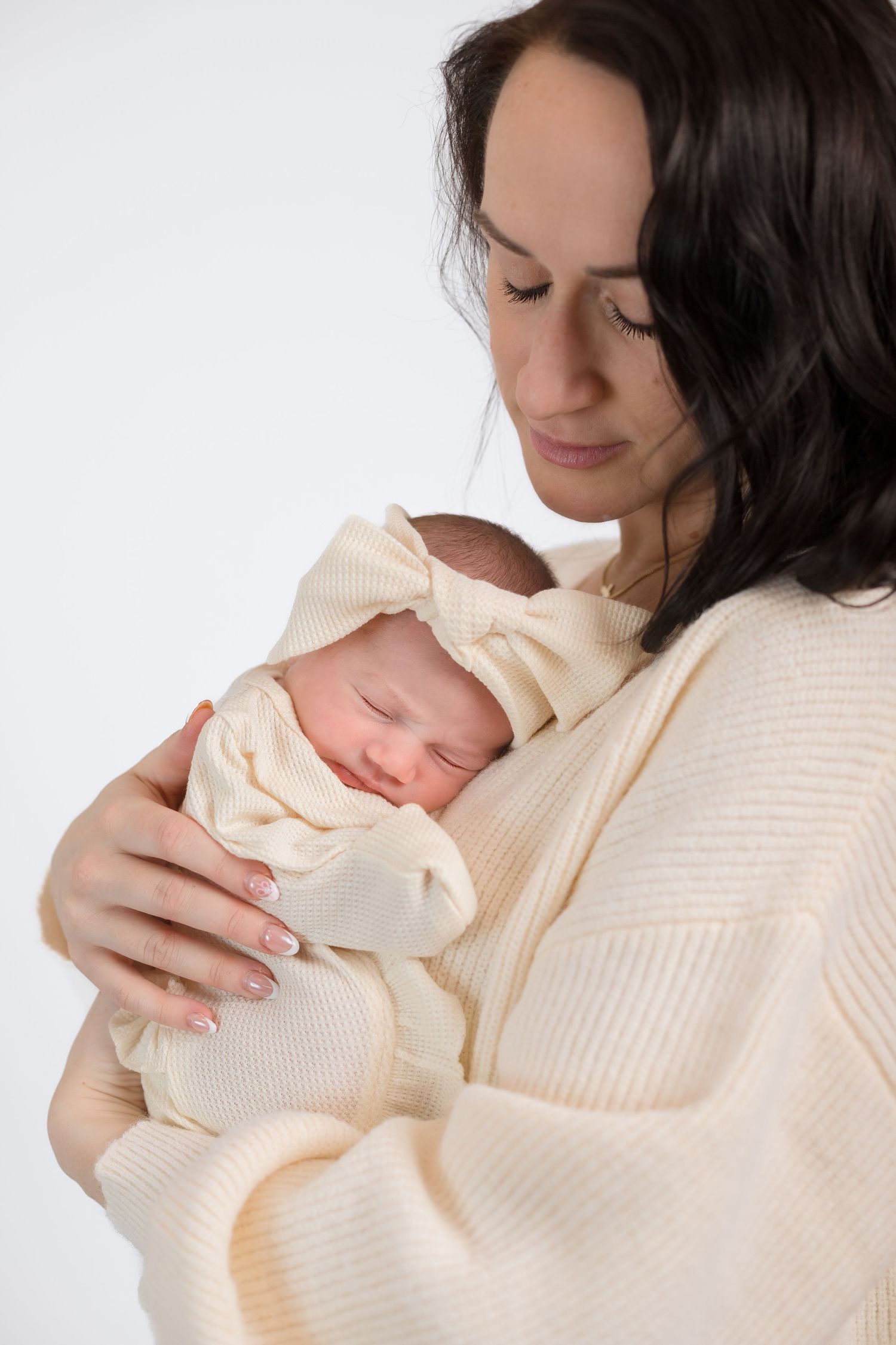 Mother in cream sweater holding sleeping newborn against white background.
