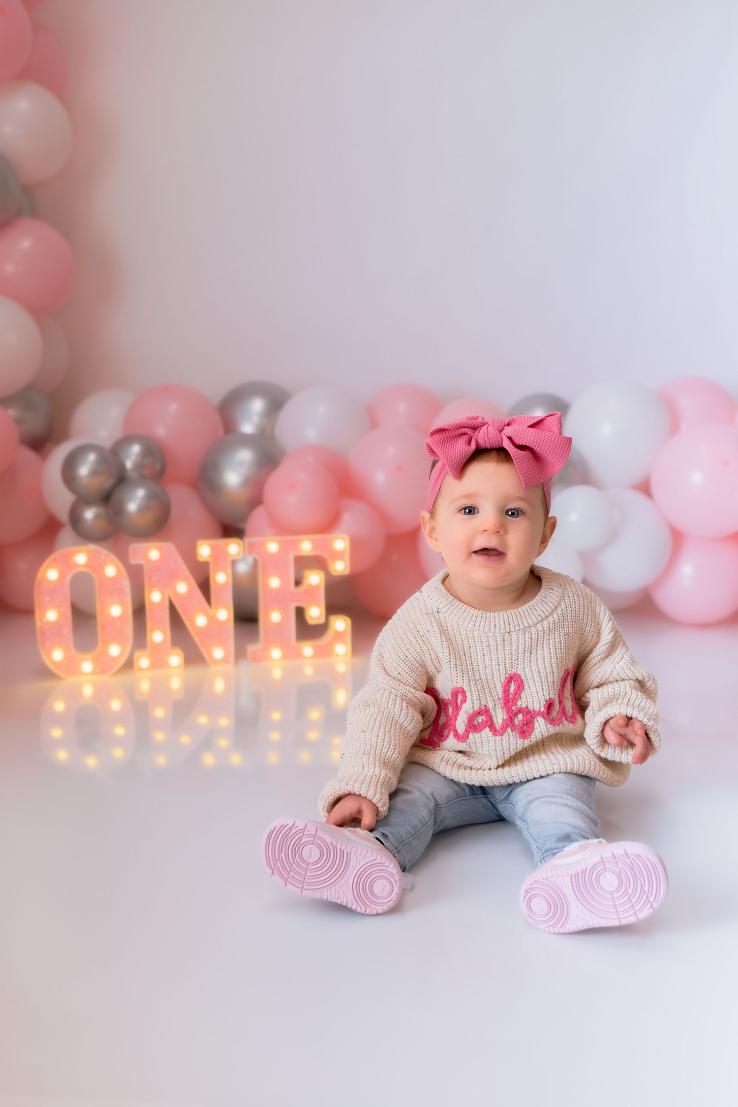 Baby wearing cream sweater and pink socks sitting by illuminated ONE sign with balloon decorations.