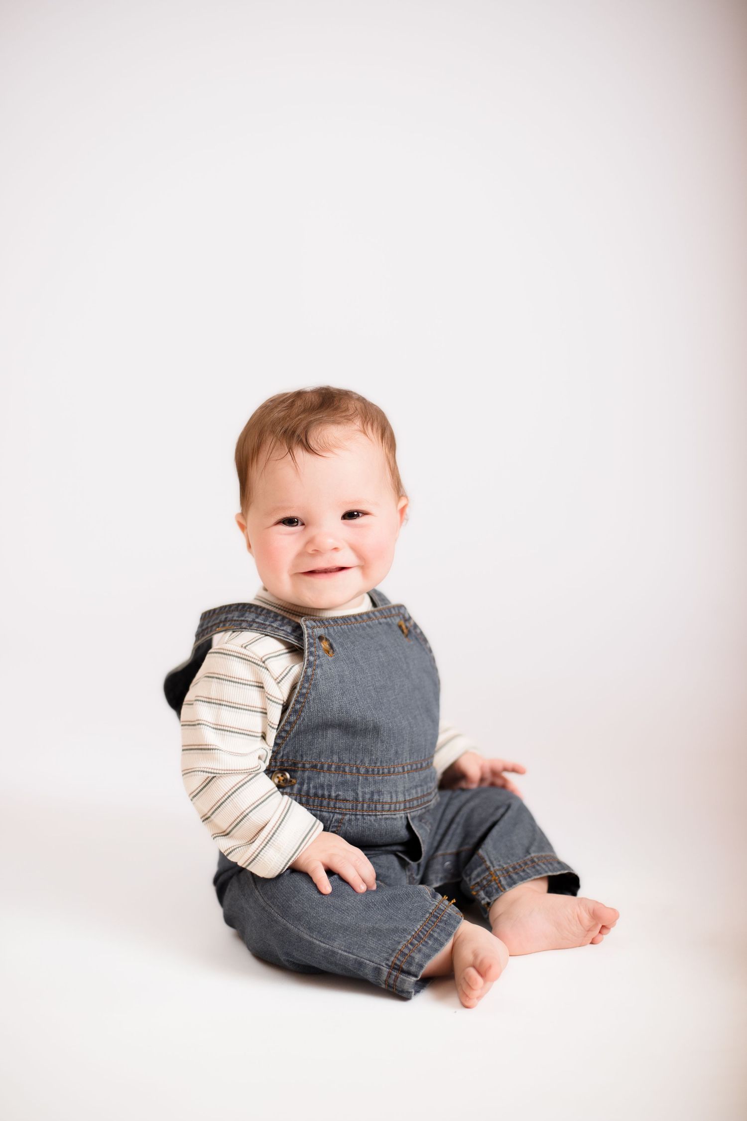 Baby wearing denim overalls and striped shirt sits on white background.