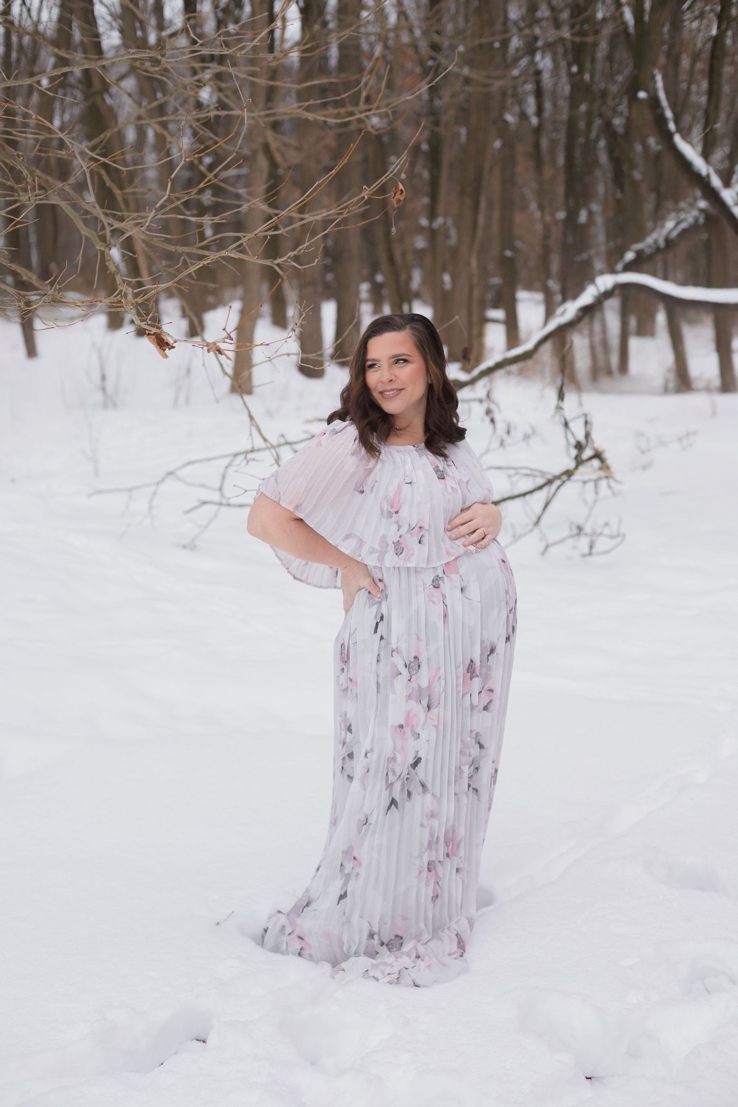 Winter wonderland maternity session featuring floral dress against snowy forest backdrop.