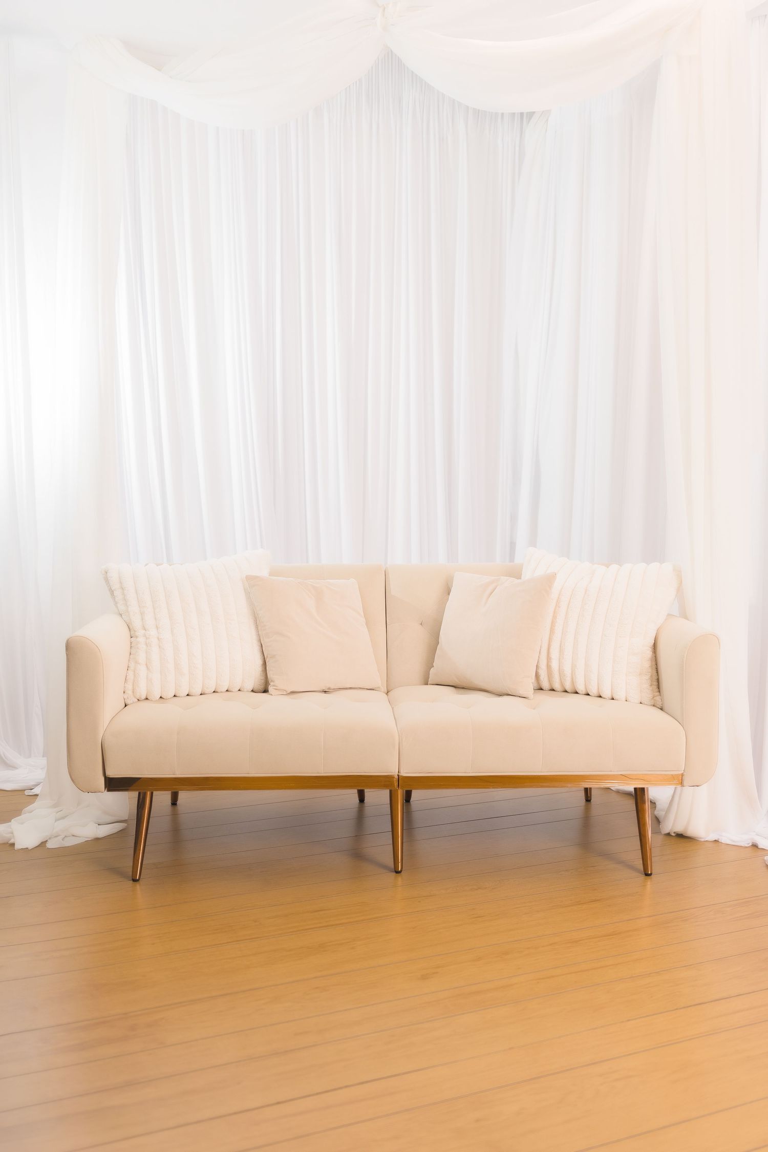 Modern beige loveseat with metal legs sits against white draped curtain backdrop on wooden floor in minimal setting.