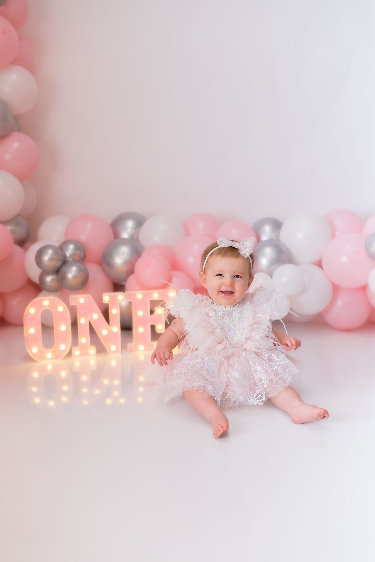 Baby in lace dress posing with pink and silver balloon arch and lit 'ONE' sign.