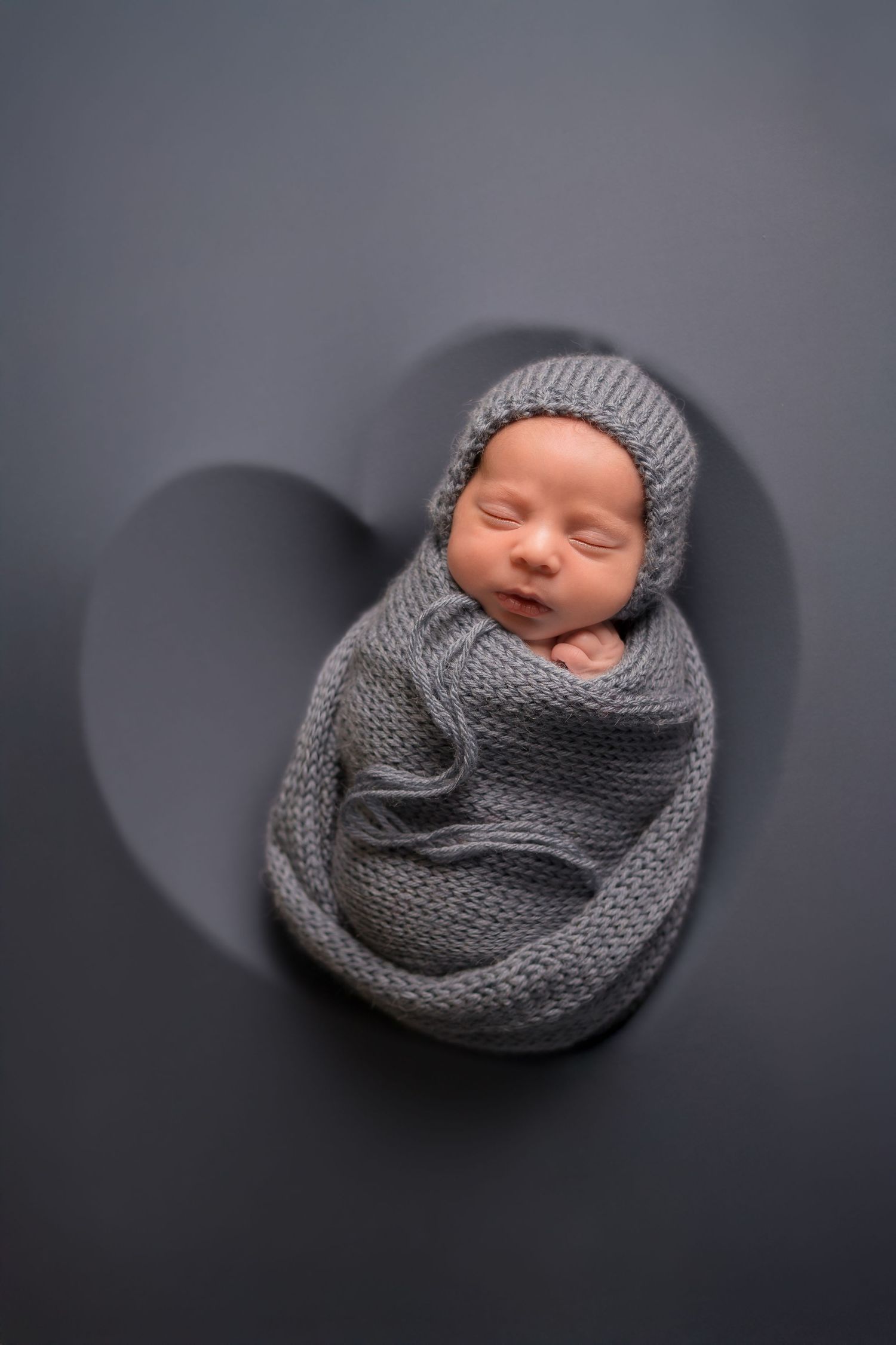 Baby sleeping peacefully while swaddled in gray knitted wrap on dark backdrop.