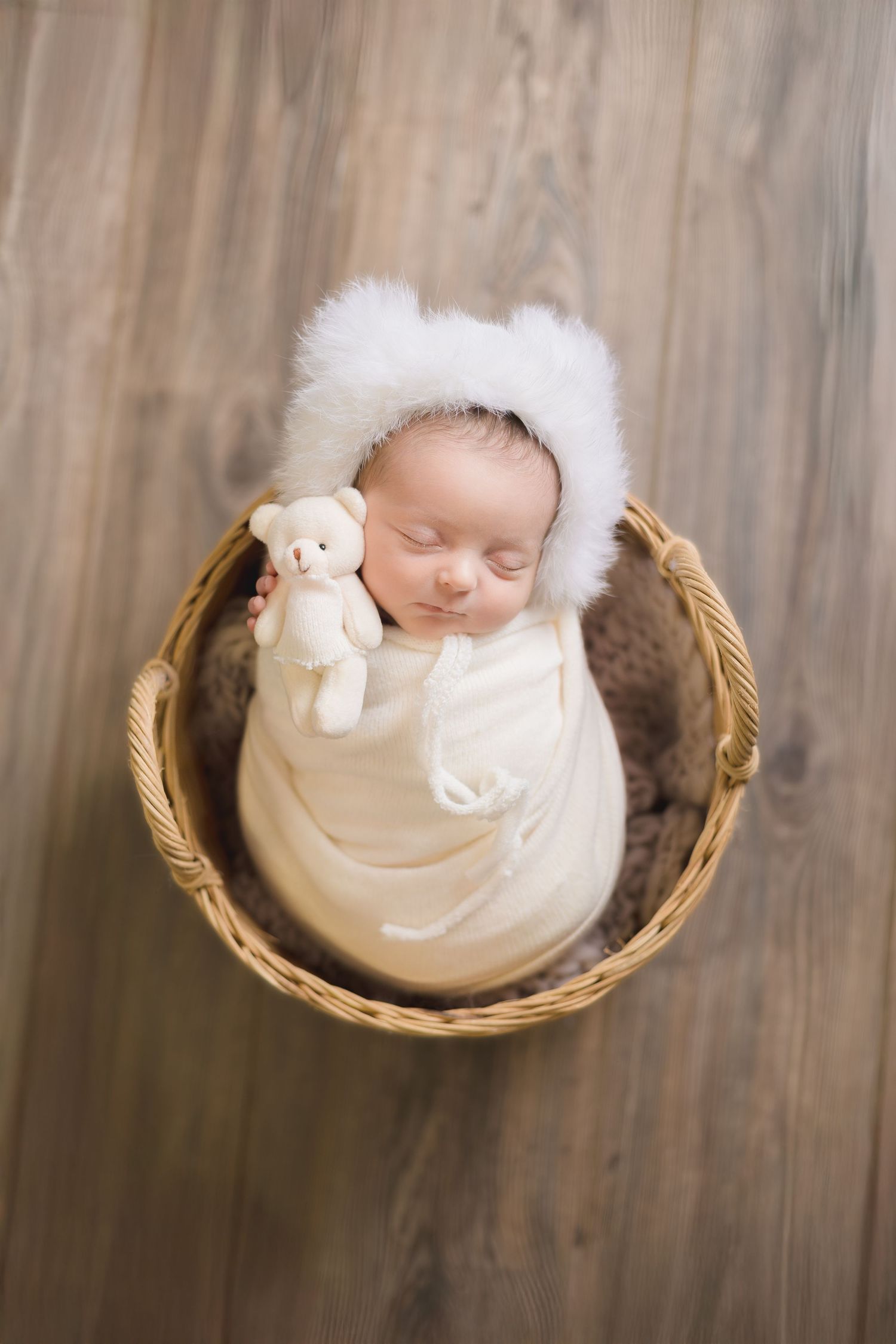A sleeping newborn baby wrapped in white fabric lies peacefully in a wicker basket with a small stuffed animal.