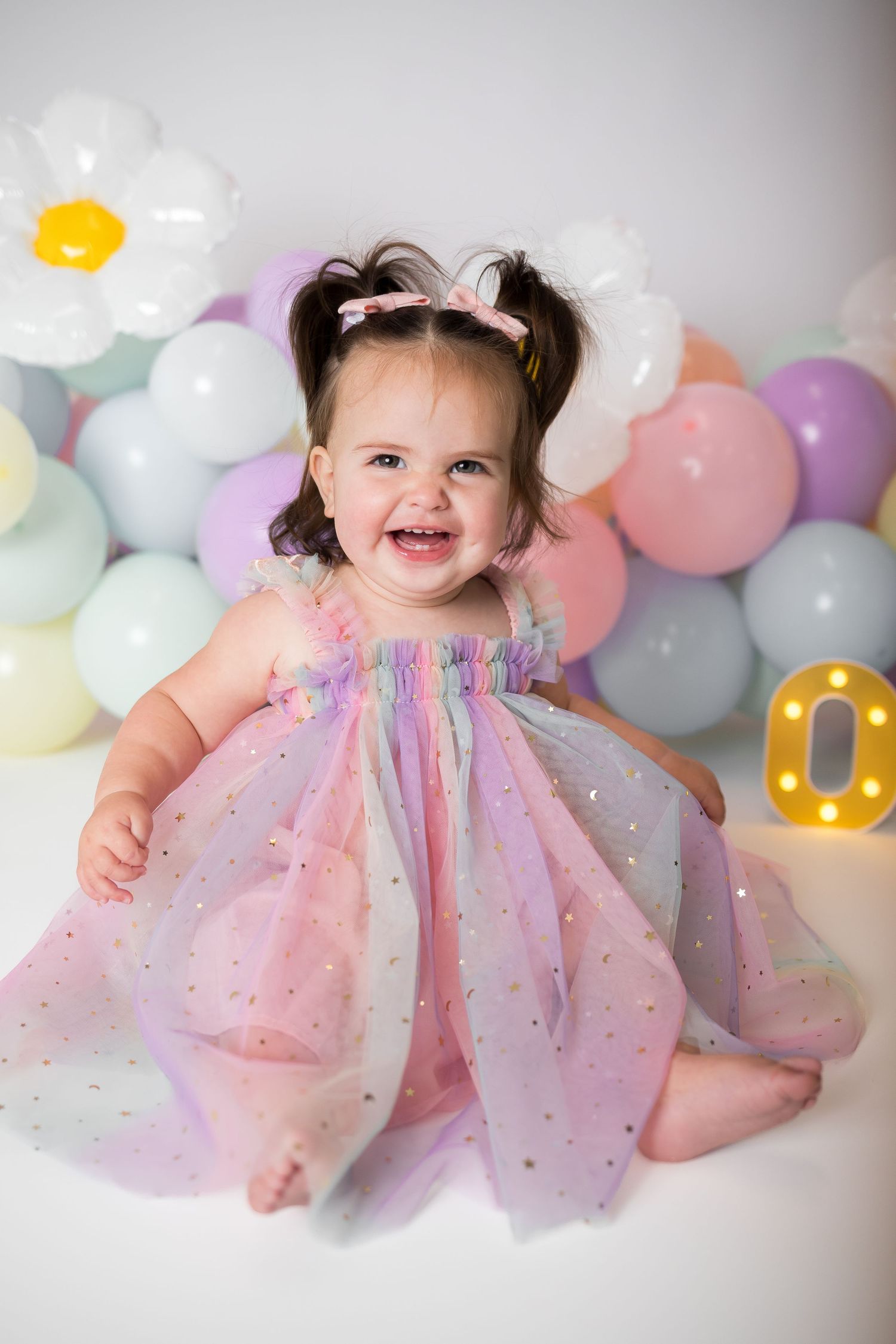 Baby in sparkly pastel dress surrounded by soft colored balloons during celebration.