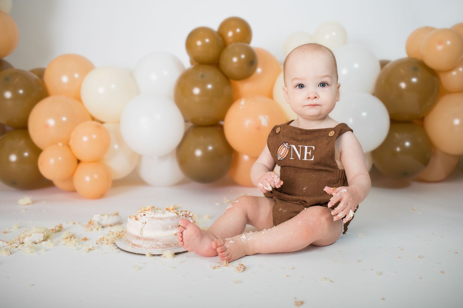Baby in brown outfit with balloon garland backdrop during cake smash session.