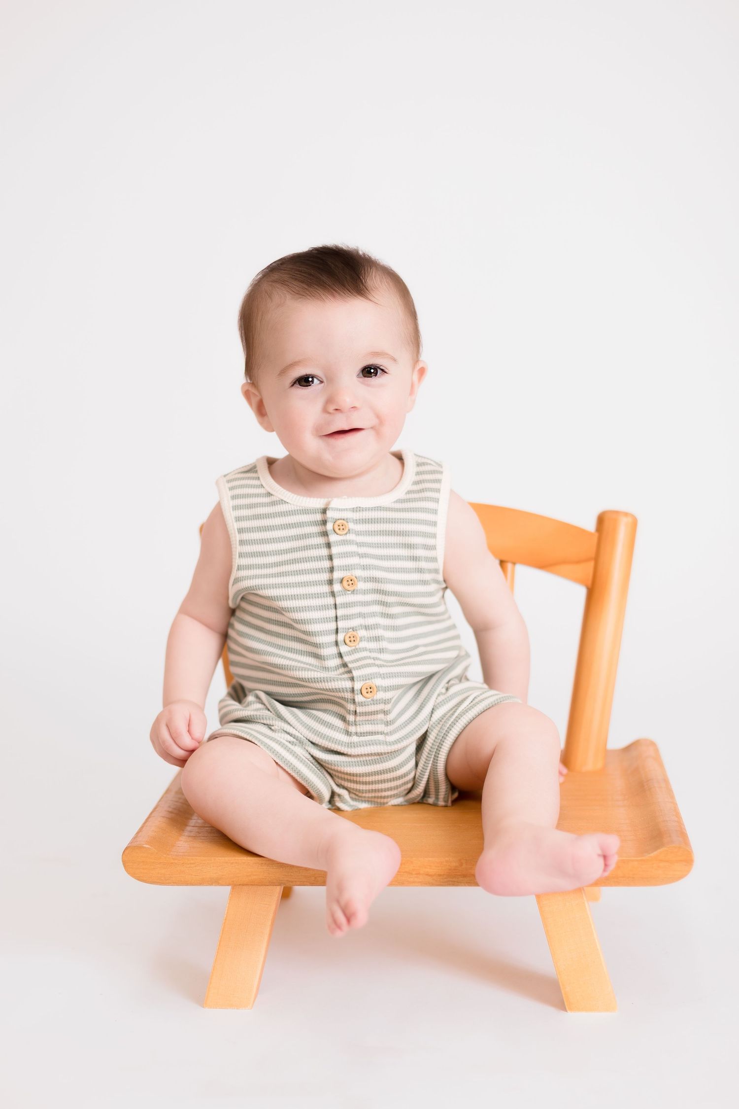 Baby in striped romper sits on small wooden bench against white backdrop.