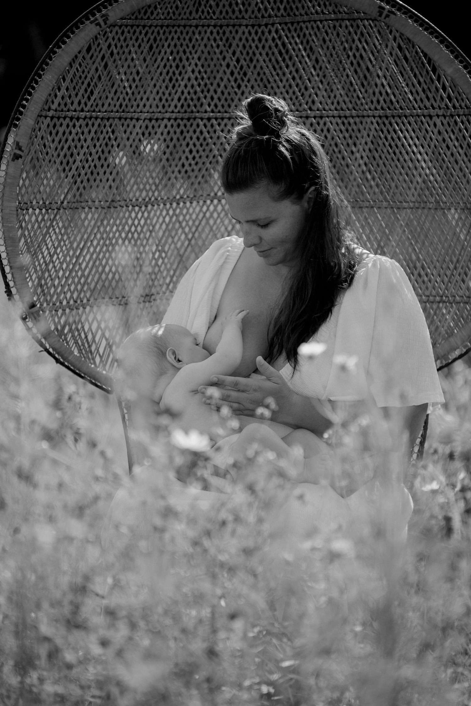 A tender breastfeeding moment captured in black and white with a woven peacock chair backdrop in a dreamy outdoor setting.