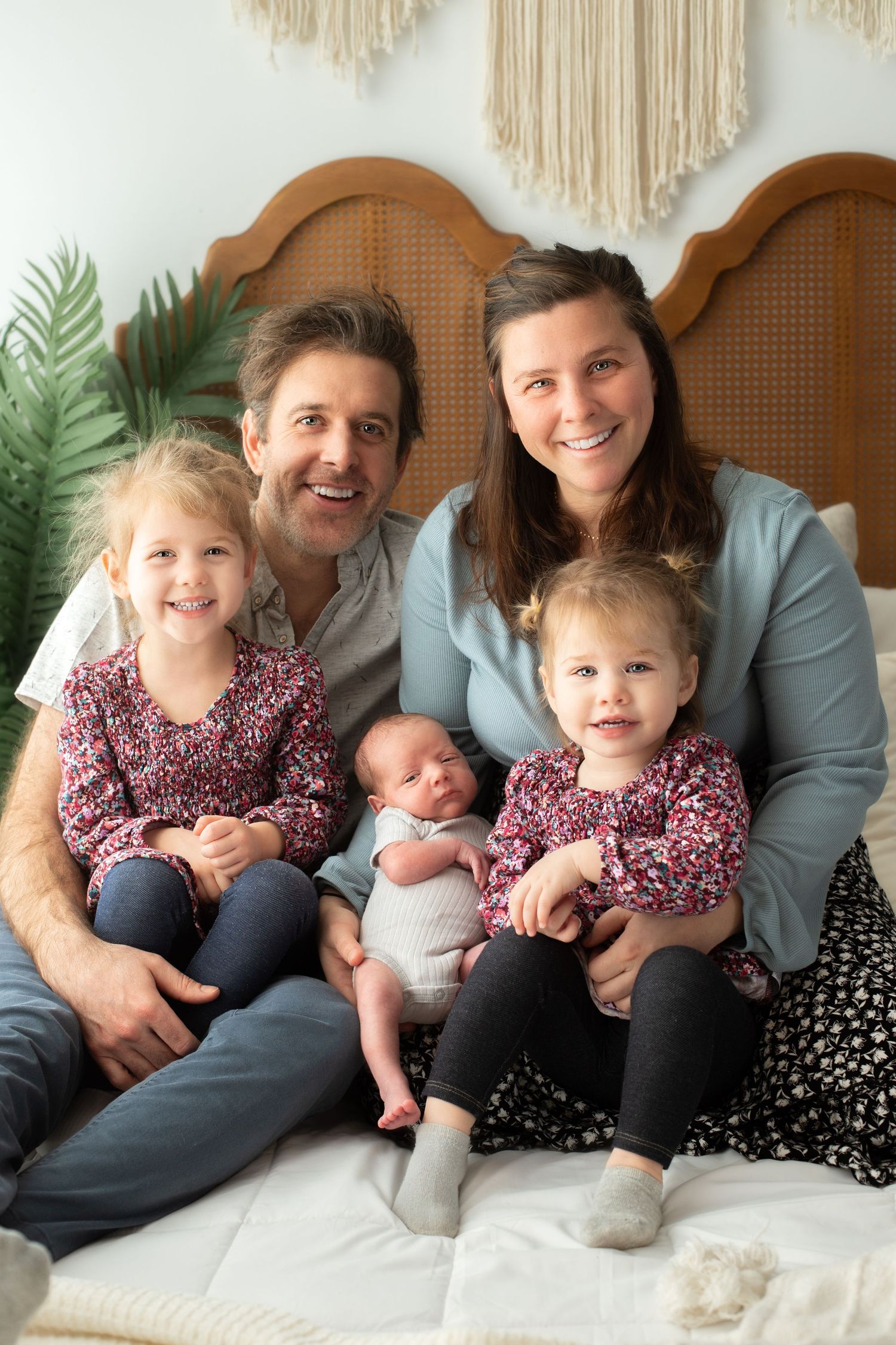Family portrait on white bed with macrame wall hanging and greenery decor.