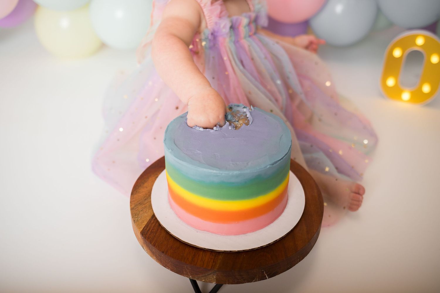 A colorful rainbow layered cake with purple frosting on top sits on a wooden cake stand.