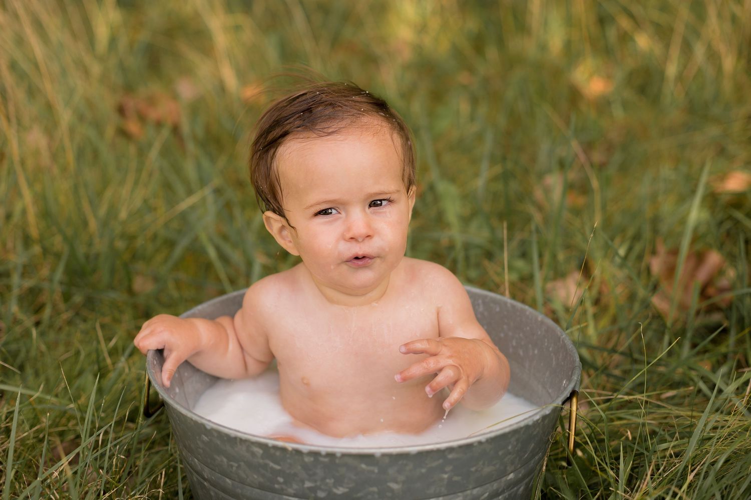 Baby sits in a galvanized metal tub outdoors surrounded by green grass in a natural setting.