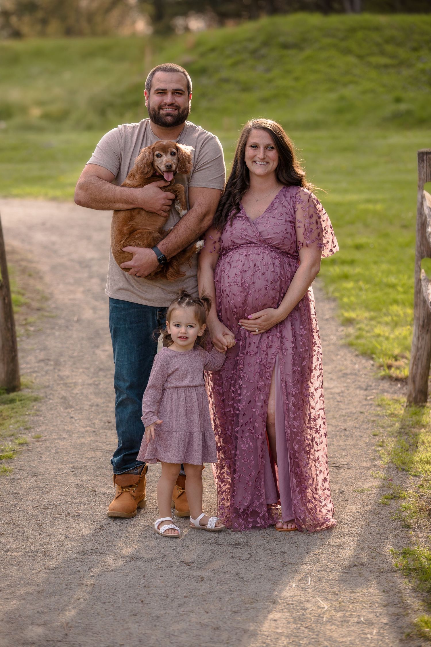 A family poses on a nature trail with their small dog, the woman wearing a flowing purple maternity dress.