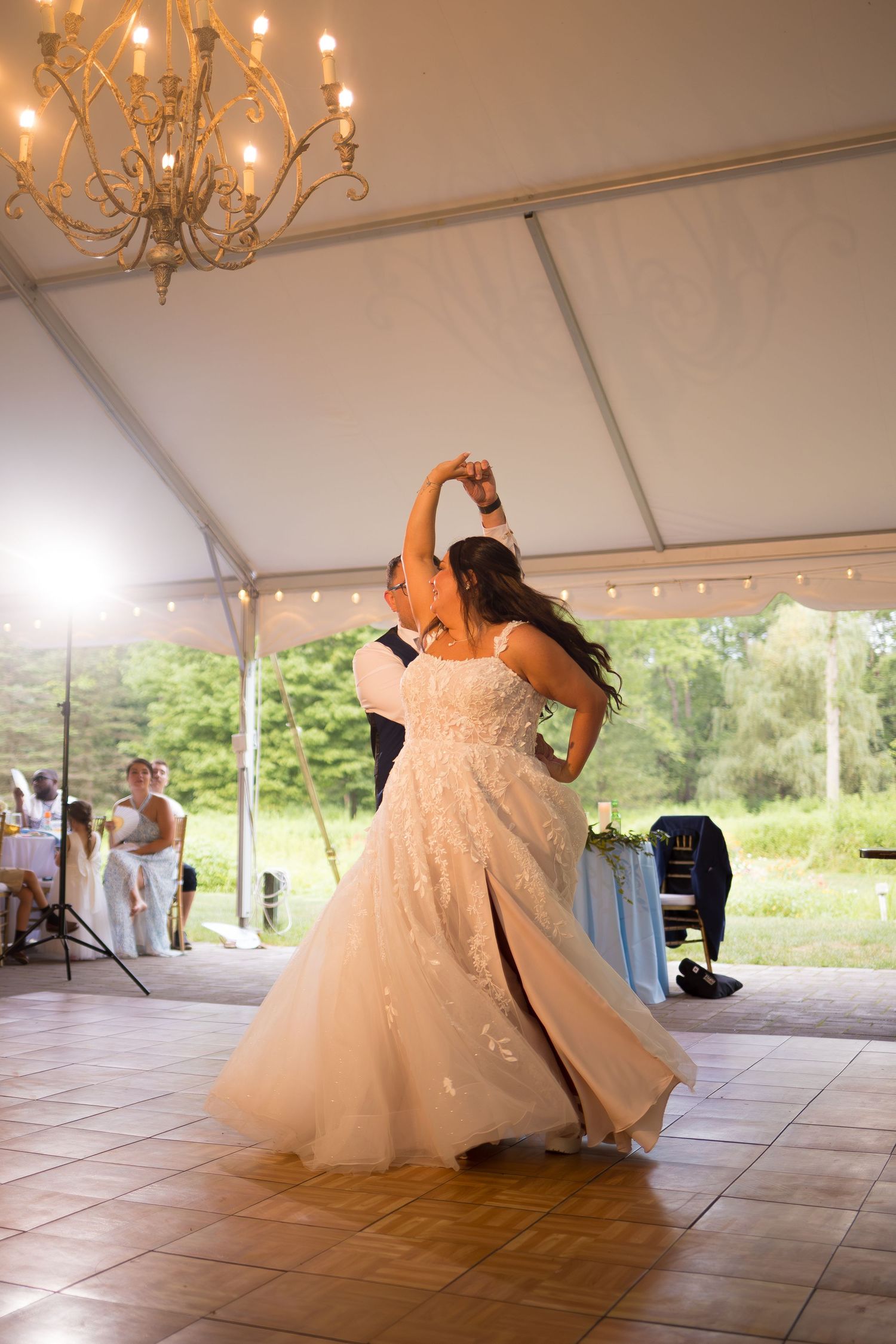 A couple performs an elegant dip during their first dance at a wedding reception under a white tent with chandeliers.