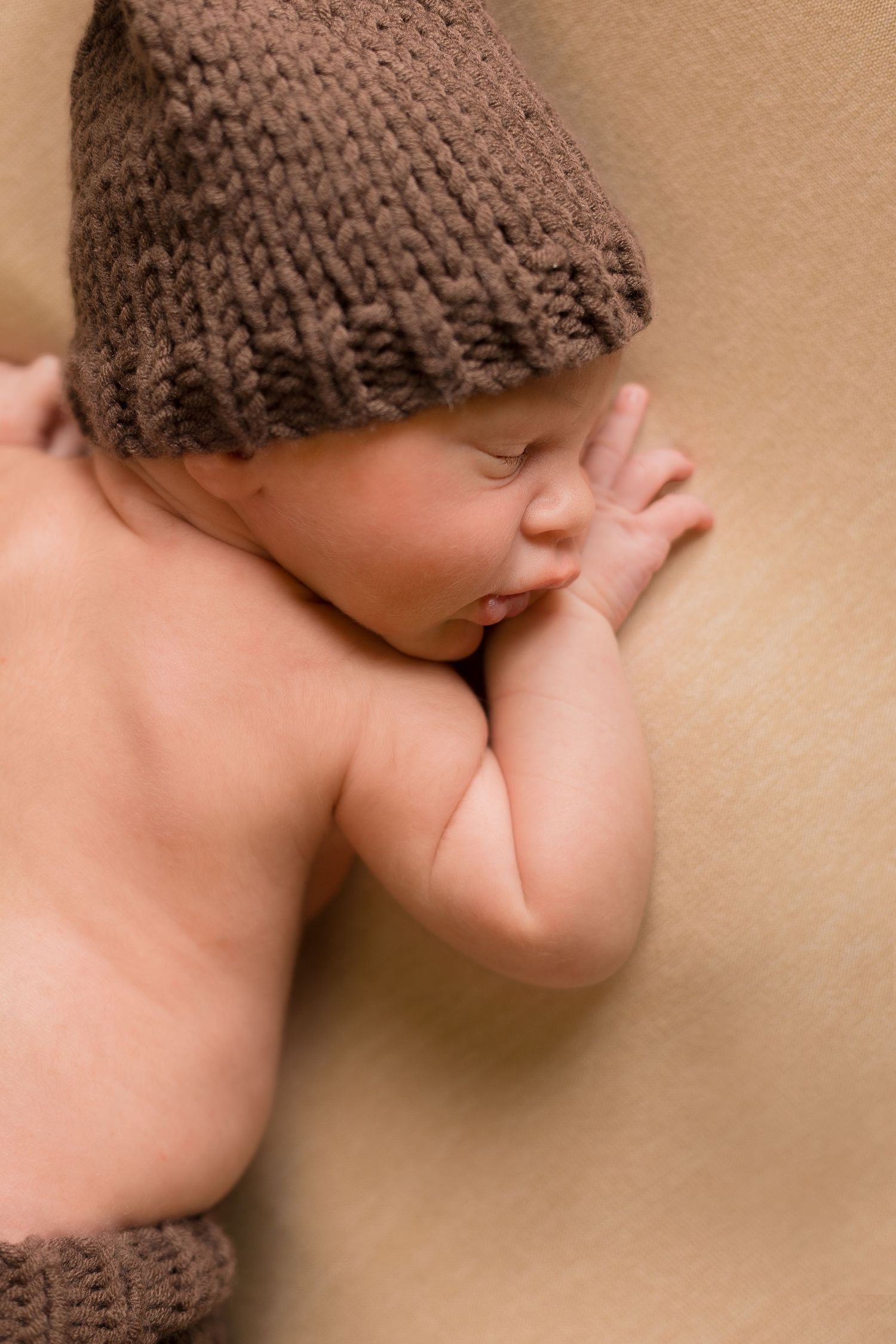 Sleeping newborn baby wearing a cozy brown knitted hat, curled up peacefully on a soft tan background.