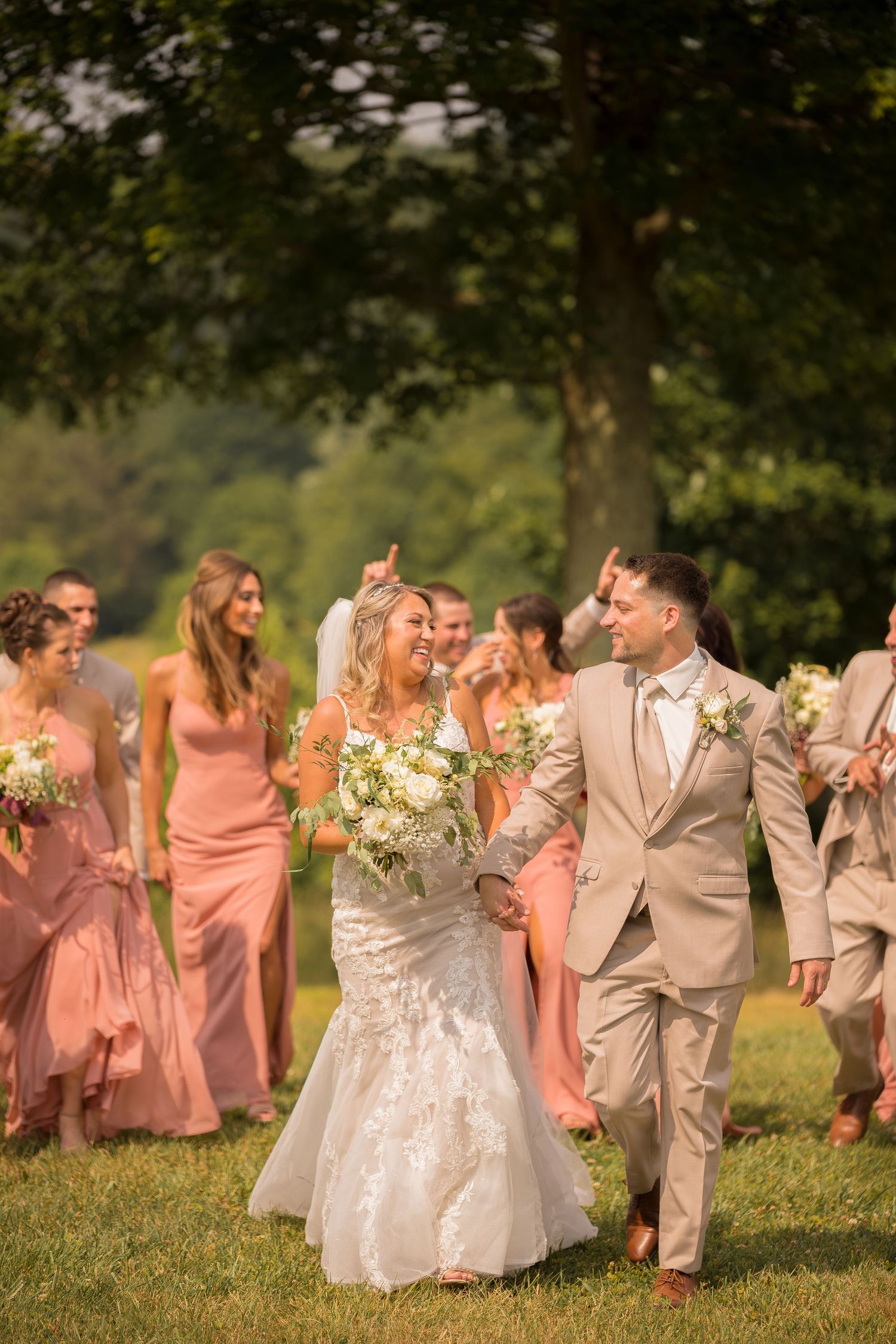 Wedding party in blush pink dresses and tan suits walking outdoors on a sunny day.