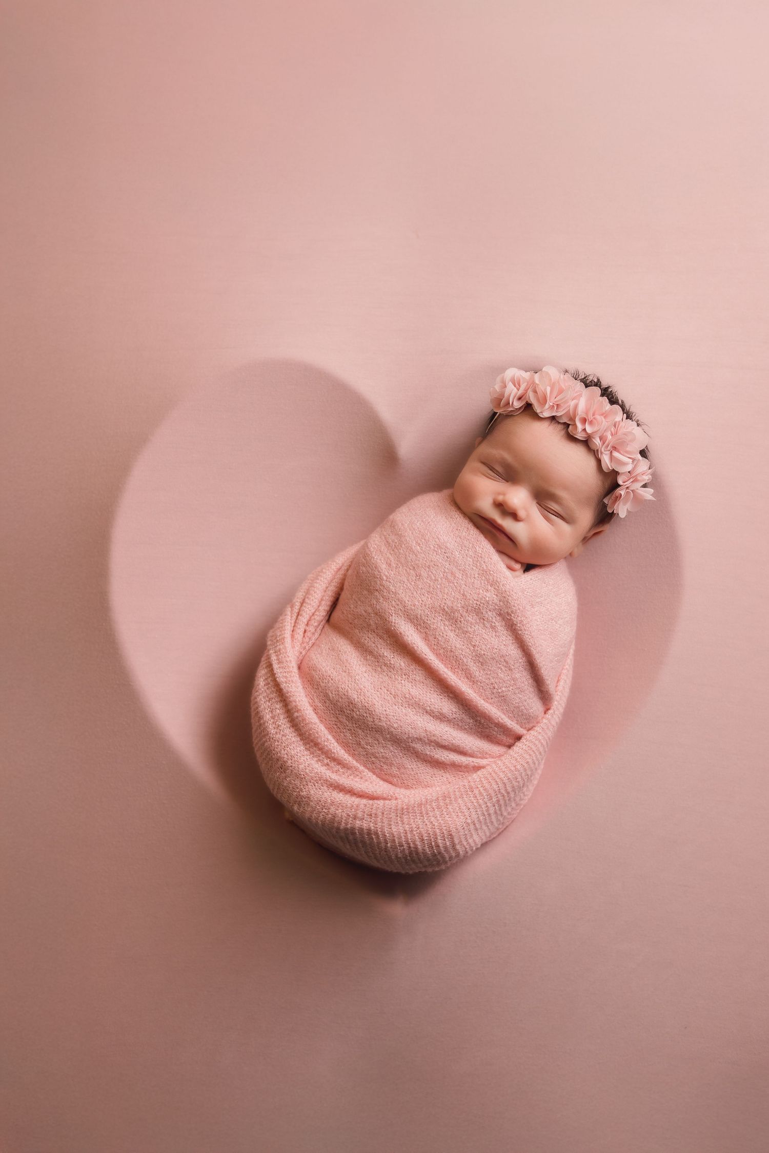 Newborn baby wrapped in pink blanket sleeping peacefully on pink background.