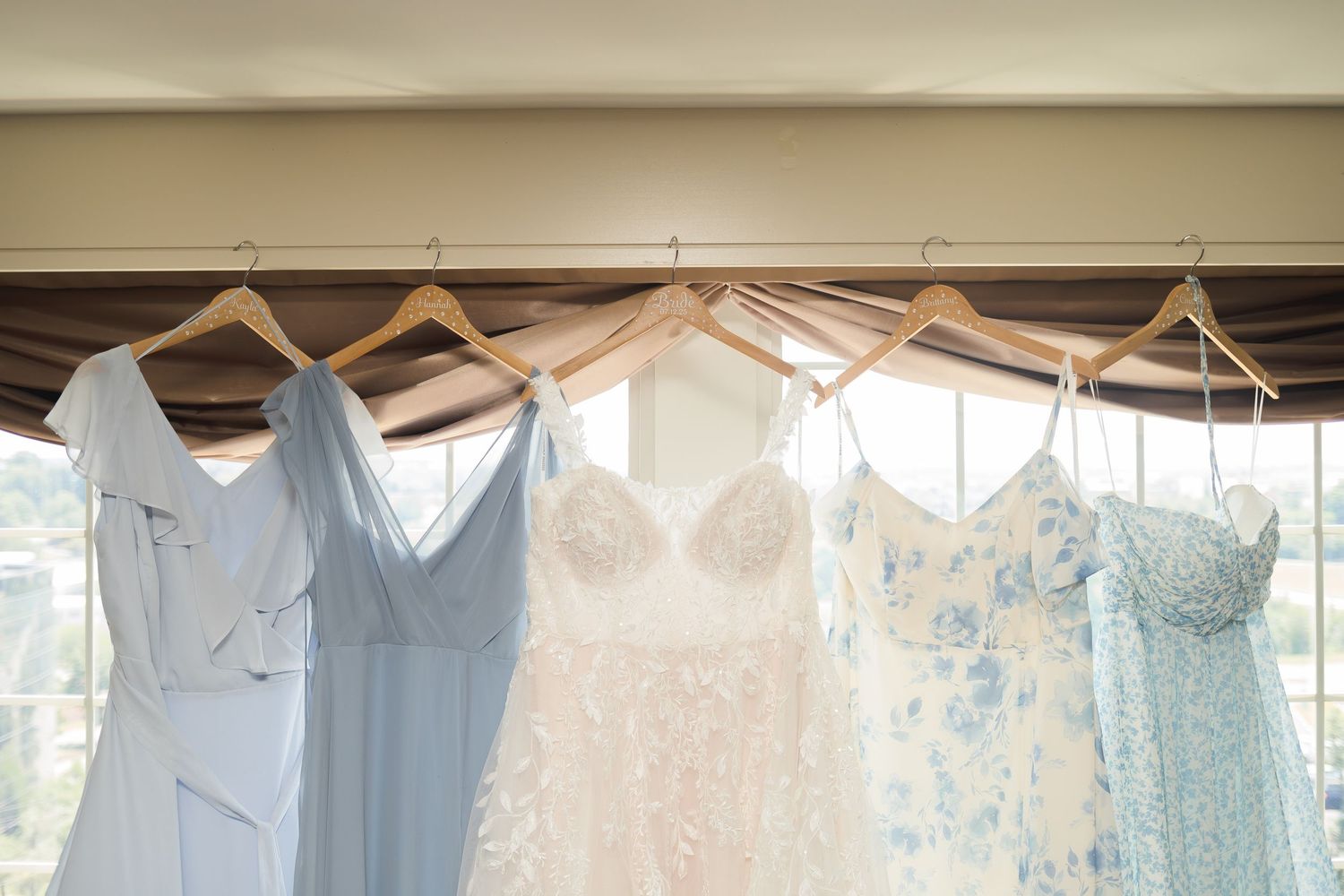 Row of pastel bridesmaid dresses and white wedding gown hanging on wooden hangers.