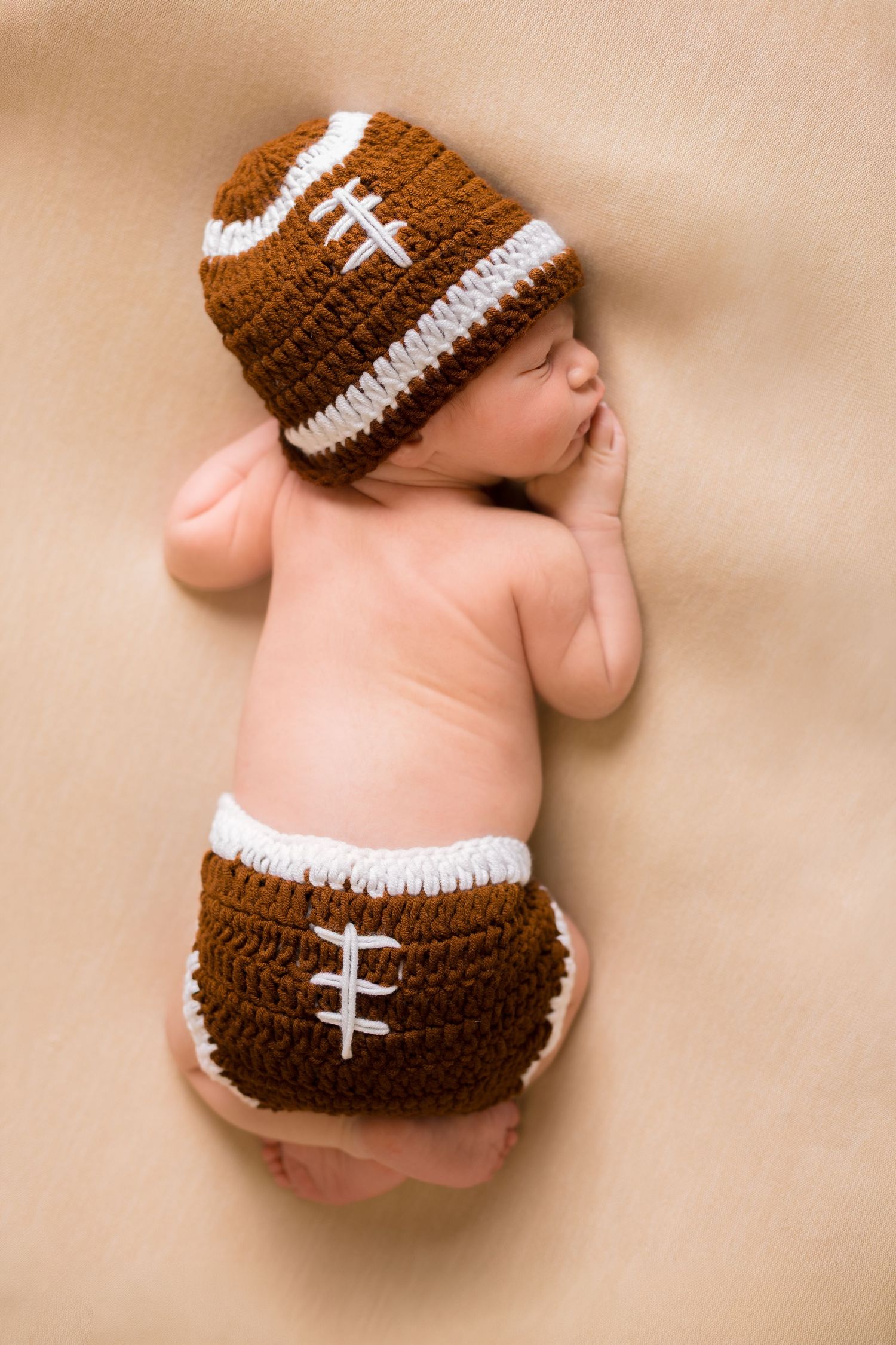 Newborn baby sleeping peacefully wearing a crocheted brown and white football hat and diaper cover outfit.
