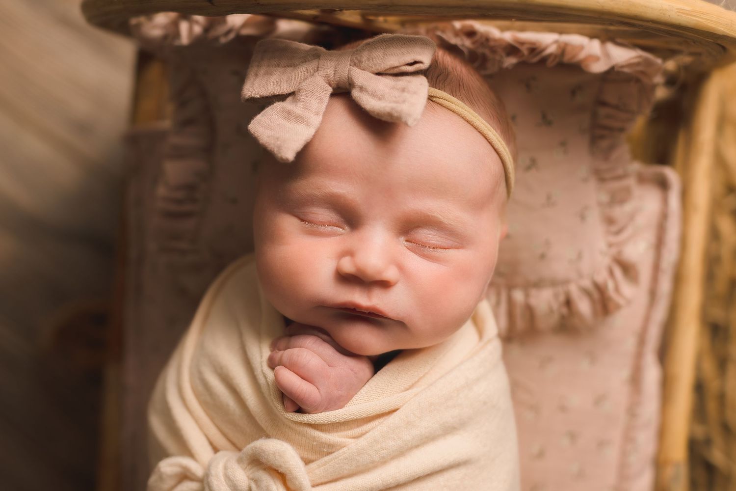 Newborn baby girl swaddled in cream wrap with pink bow headband sleeping peacefully in a wooden basket.