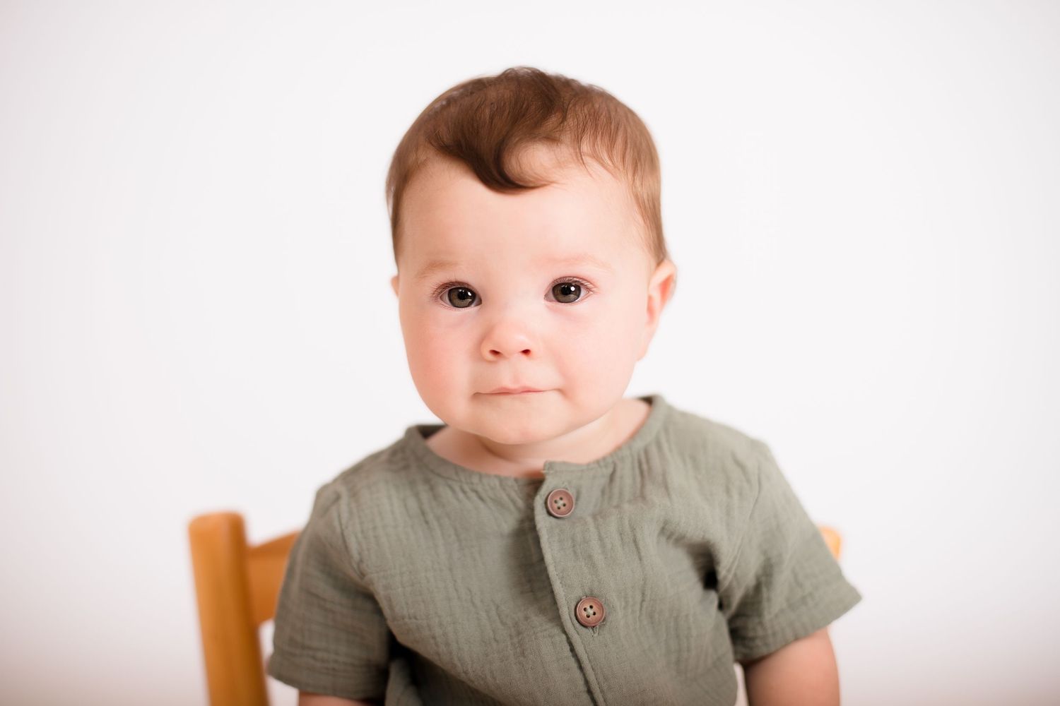 Baby in olive green outfit sits on wooden bench against white background.