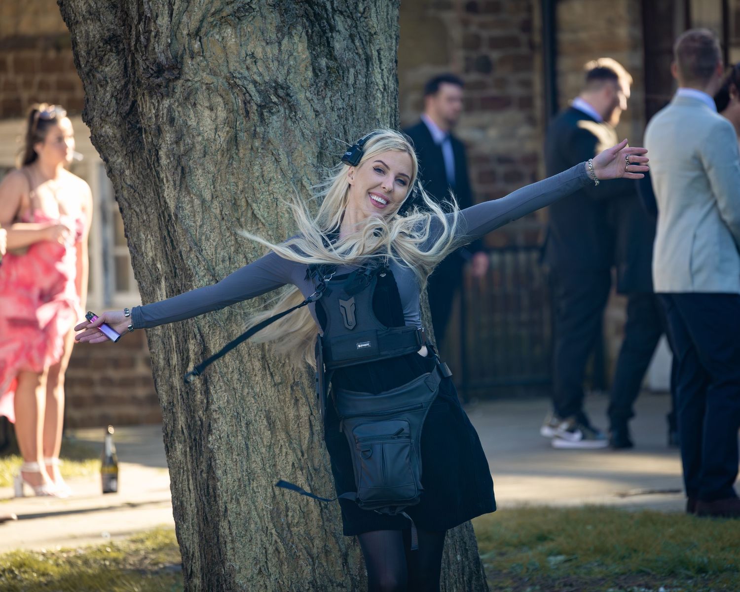 Energetic blonde woman hugging a tree at an outdoor event while carrying camera equipment. Hertfordshire, Cambridgeshire, UK
