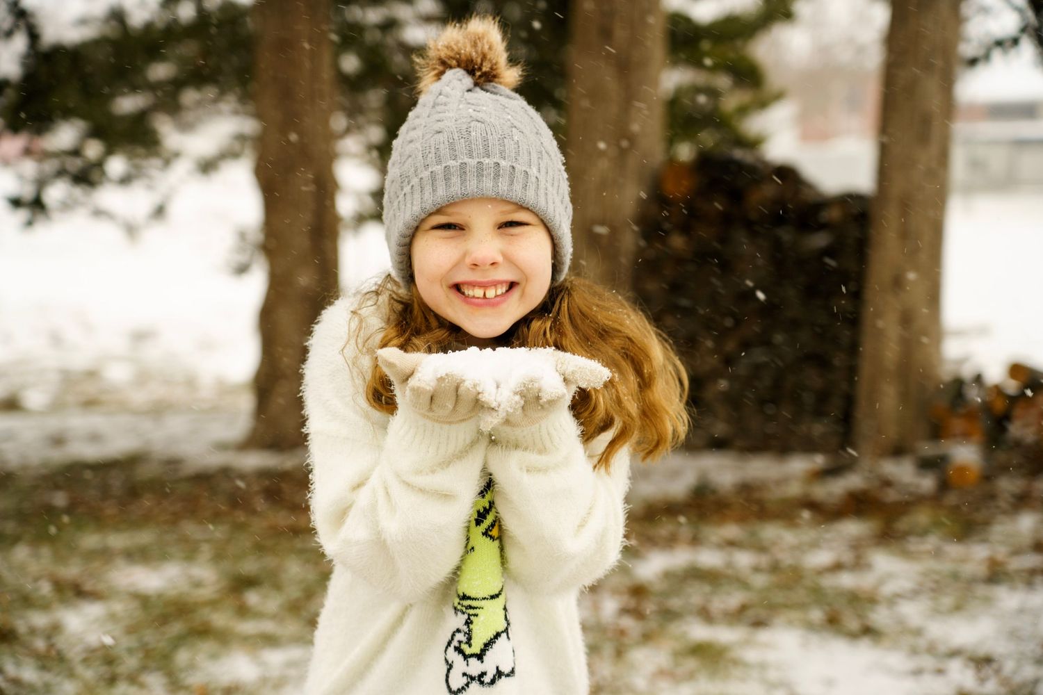 A smiling figure in a white winter coat and knit beanie poses playfully in the snowy outdoors.