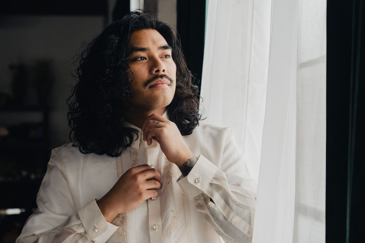 A person with curly hair wearing a white shirt poses thoughtfully by a window in moody lighting.