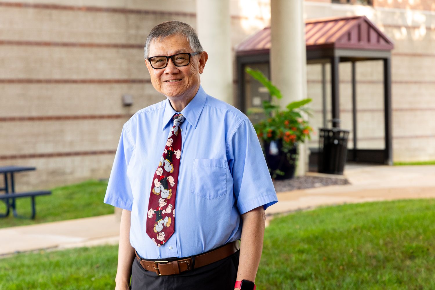 Professional portrait in a light blue dress shirt and burgundy tie against a stone building backdrop.