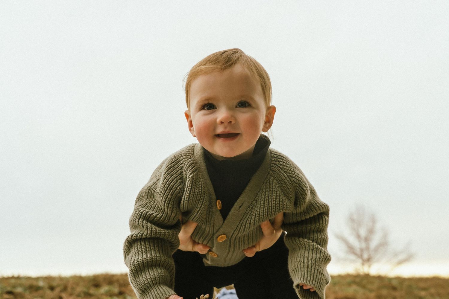 A small child in a green sweater smiles while playing outdoors in natural light.