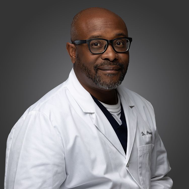 Medical professional in white coat and glasses poses for studio portrait against dark backdrop.