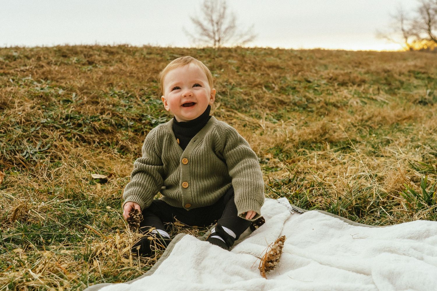A child in a brown sweater sits on a white blanket in a grassy field during autumn.