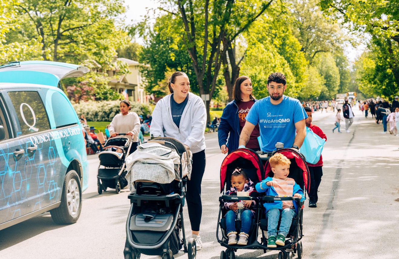 People walking and pushing strollers along a tree-lined path on a sunny day.