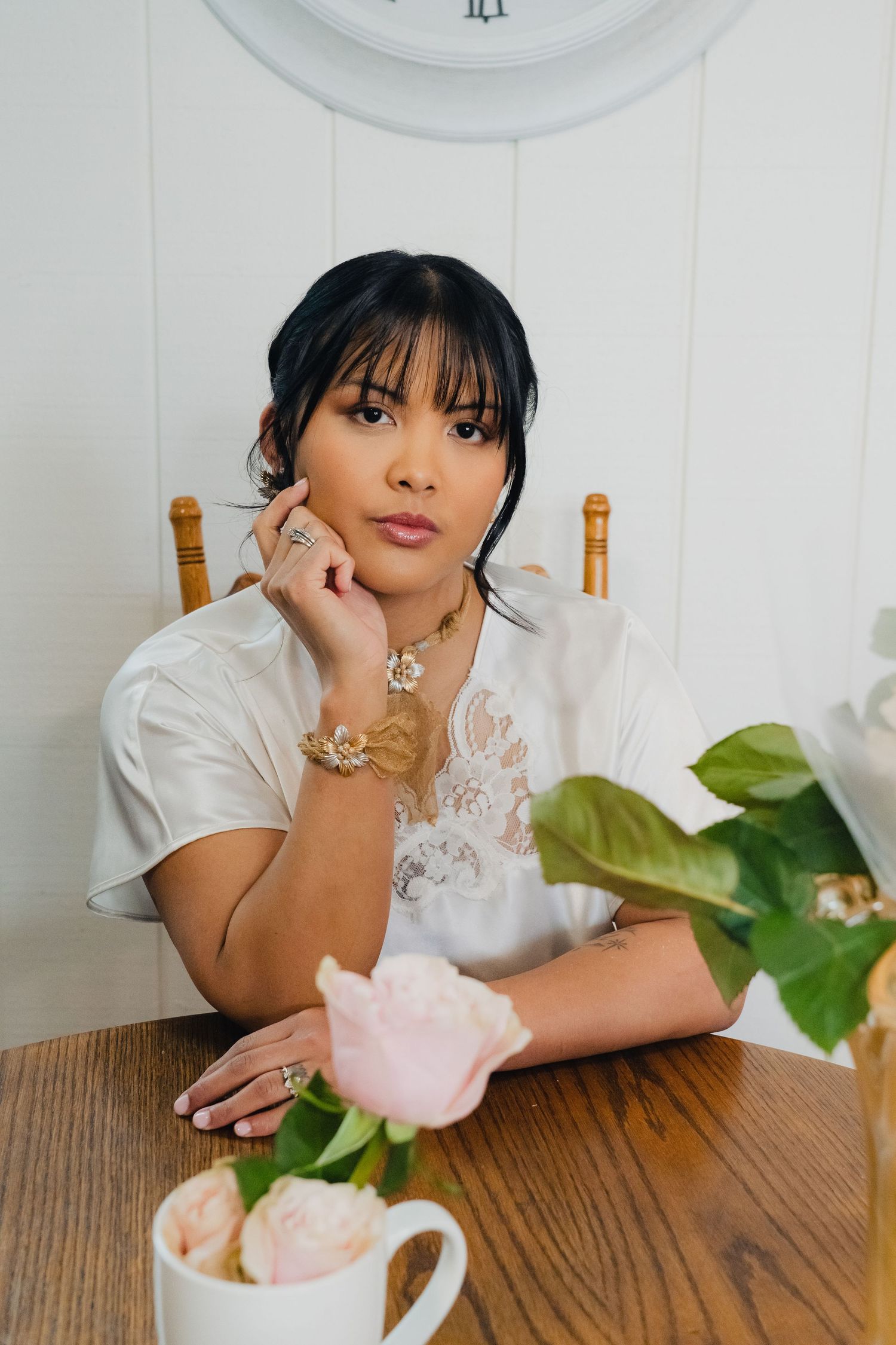 A pink rose in a glass vase sits on a wooden table with white paneled walls and a clock in the background.
