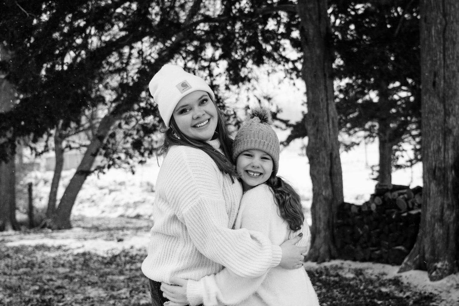 Black and white winter portrait of two people embracing outdoors while wearing cozy white sweaters and knit hats.