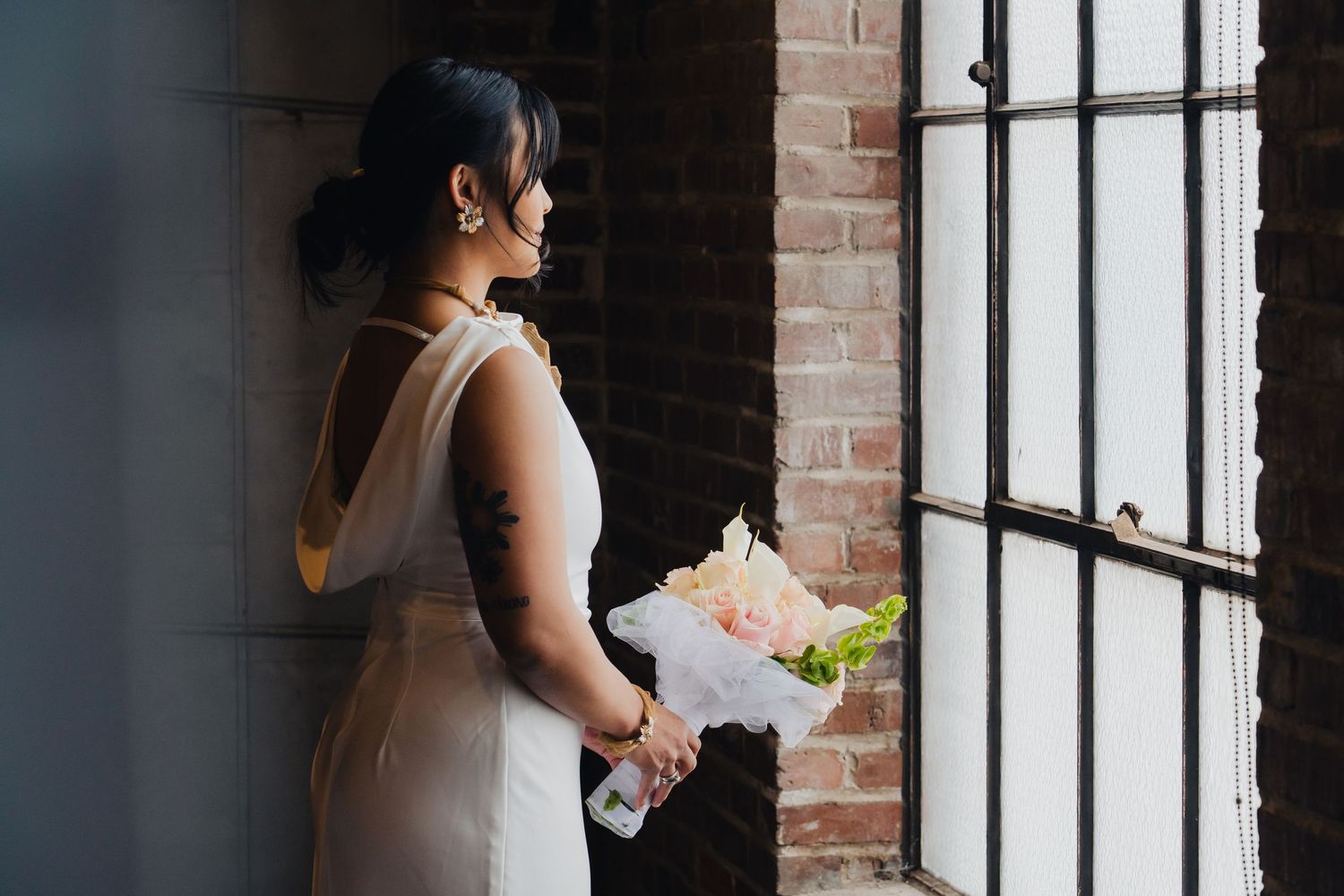A person in a white dress holds a bouquet of white flowers while standing near a brick wall and window.